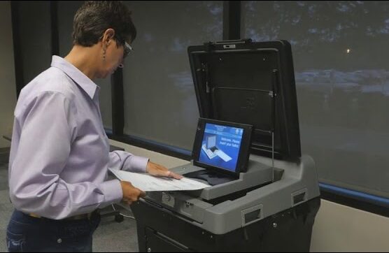 DS200 Election worker wearing lavender shirt and jeans demonstrates how to insert ballot into gray scanner on black base. Scanner has blue screen that reads "Welcome, please insert your ballot."