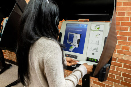 Voter with long, straight black hair and gray sweater inserts ballot into ExpressVote voting machine