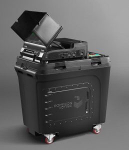 A black container positioned on wheels is displayed. A large door with the words “Dominion Voting” is on the side. Atop the container is a black base, including a scanner, and an upward-angled black plastic privacy partition surrounding an LCD screen.