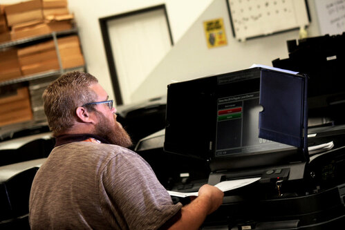 White man with glasses and red beard wearing a purple shirt holds a ballot up to a black ballot scanner slot below a black screen with red and green buttons.
