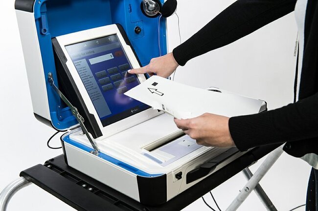 Verity_Scan_650 White ballot scanner with blue touch screen and blue case rests atop black platform. White hand of person wearing black shirt touches screen. Screen has number buttons and reads "Ready for voting."